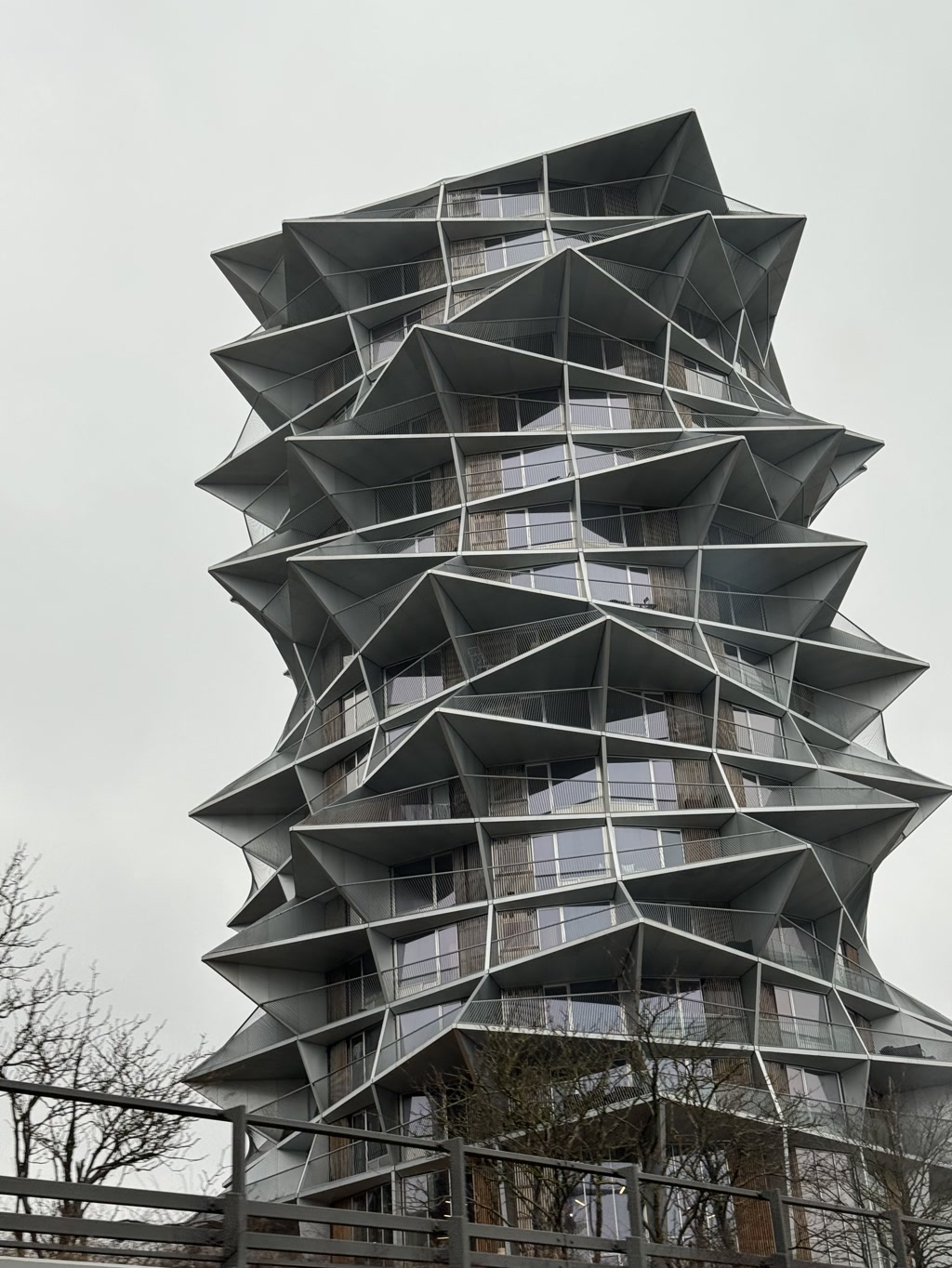 A modern multi-story building displaying a distinctive architectural design with a series of repeating geometric shapes that appear to be stackable modules. Each module consists of a star-like shape with protruding corners, creating a sense of depth and dynamic form. The facade is dominated by gray hues, with balconies enclosed by metal railings that complement the avant-garde structure. The sky is overcast, and no people are visible in the view, which also includes a glimpse of a wooden fence and bare tree branches in the foreground.
