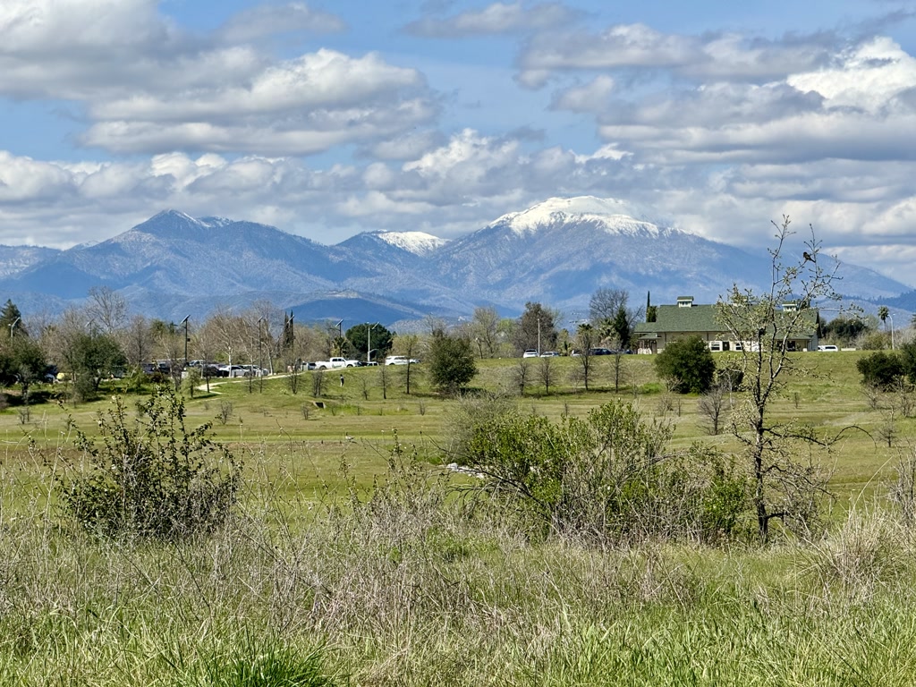 A scenic view of a verdant field with occasional shrubs under a partly cloudy sky. In the middle distance, there's a paved area with several cars parked, near what appears to be a large, two-story green-roofed building. The background is dominated by impressive mountains covered with a mix of greenery at lower altitudes and snow caps at their summits.