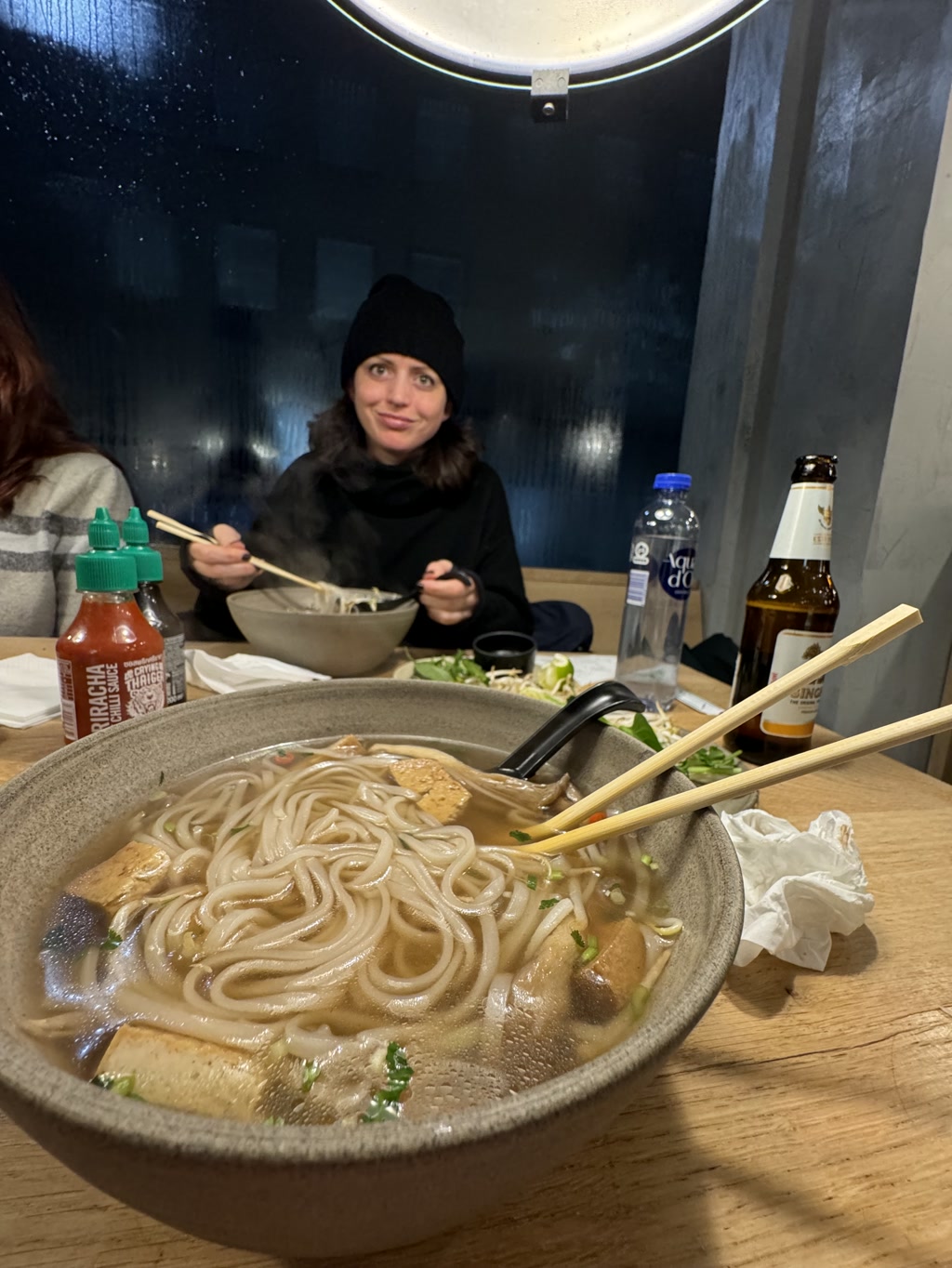 A person is sitting at a dining table in a restaurant, with a large bowl of steaming udon noodle soup in front of them. They are holding a pair of chopsticks in one hand and a spoon in the other, poised to eat. The individual appears to be wearing a black beanie and a black turtleneck. The table also has a bottle of beer and a water bottle to the right, along with a bottle of Sriracha hot sauce. The environment suggests an informal setting, possibly during evening hours as indicated by the darkness visible through the round window behind the diner.