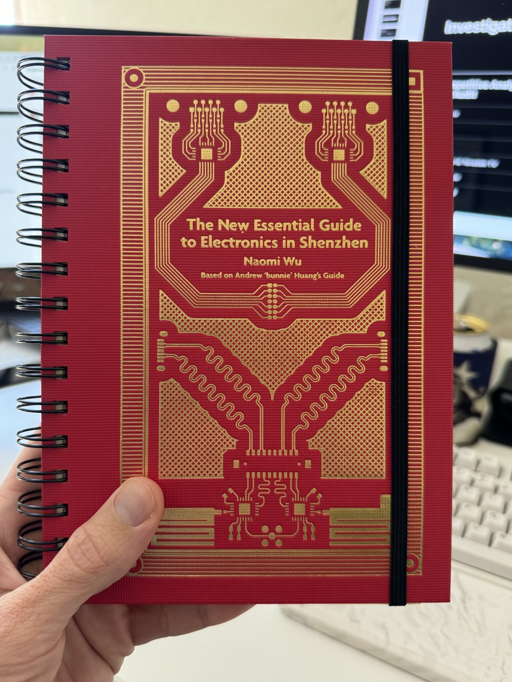 The item shown appears to be a spiral-bound notebook with a hardcover featuring a gold-yellow printed circuit board (PCB) design on a red background. The cover design features intricate circuitry patterns typical of electronic devices, suggesting a technical or educational theme to the contents of the notebook. The PCB tracks lead to a central figure evocative of an integrated circuit or microchip. The notebook has a black elastic band to keep it closed.