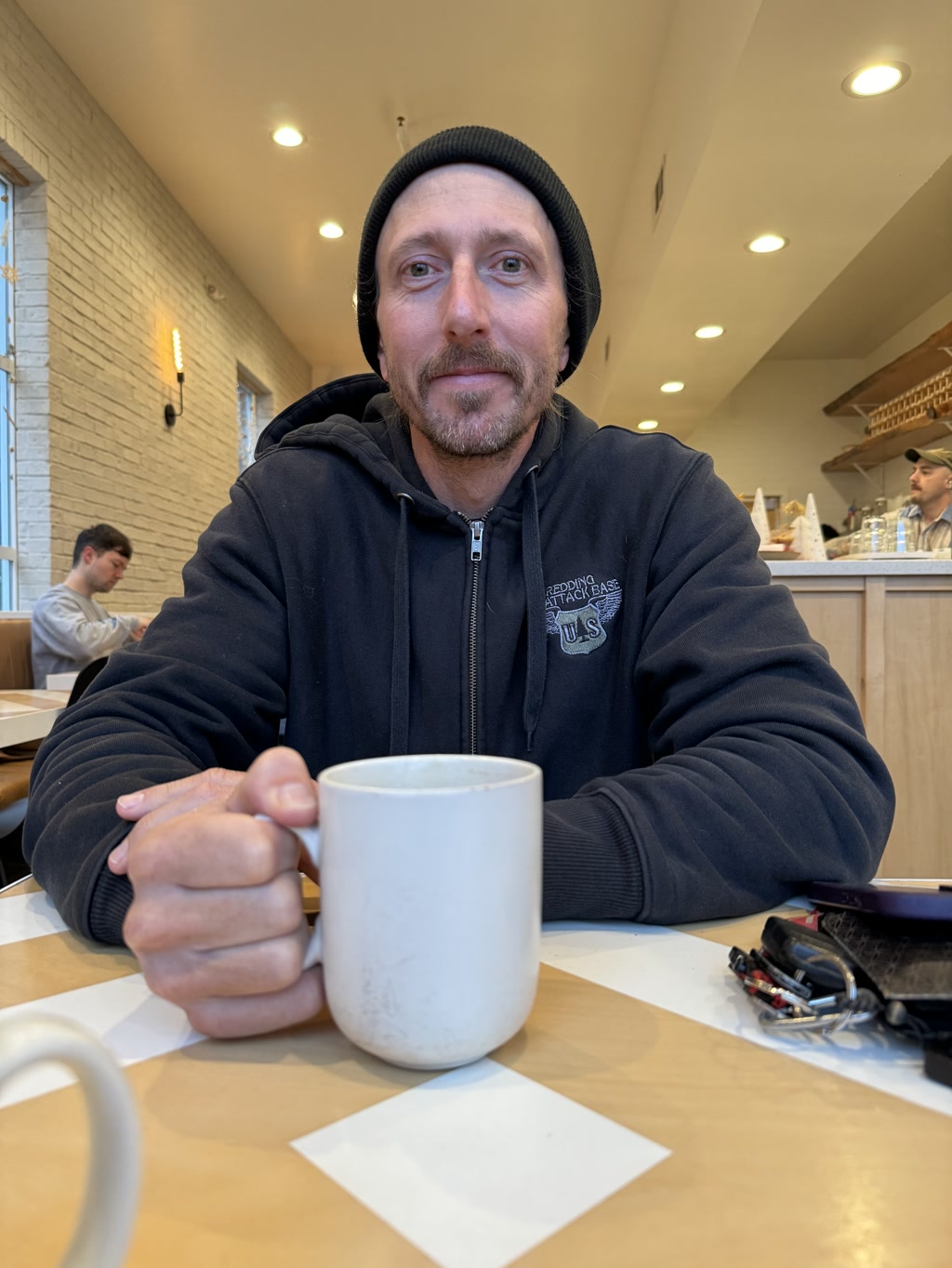 A person is sitting at a table, holding a white mug. They have a close-cut beard and mustache and are wearing a black beanie and a black hoodie with a zipper and white lettering. The hoodie has an emblem on the left side with some text and an illustration of a jumping fish that reads 'REDDING UPDATE ATTACK TEAM'. On the table there are also some keys and a smartphone with a purple case. The background suggests the setting is a cafe, with bright interior lighting, additional seating, and what appears to be a service counter.