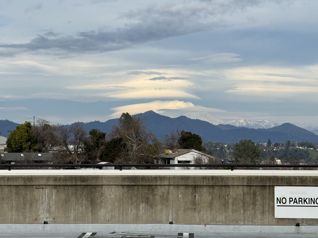 A tranquil view of a mountainous landscape with the snow-capped peaks in the distance visible under a partly cloudy sky. In the foreground, there is a concrete barrier with a sign that indicates a parking restriction.