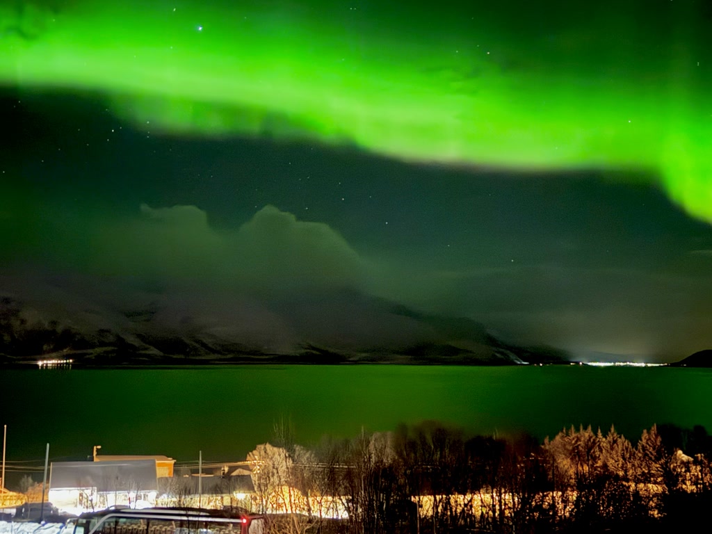 A vibrant display of the Northern Lights, or Aurora Borealis, illuminates the night sky with a bright green hue, creating an ethereal arch over a winter landscape. Below, a body of water reflects the green light, while snow-covered mountains loom in the background. The foreground shows a small neighborhood with illuminated buildings and streetlights, adding a warm glow to the cold scenery. A few trees and vehicles are visible, completing the serene winter night tableau.
