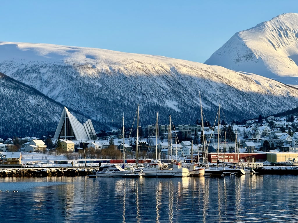 A picturesque coastal landscape showcasing a small harbor filled with various sizes of boats moored along a jetty. The foreground is dominated by calm, reflective waters. In the background, there stands a distinctive triangular-shaped building that appears to be a modern structure, such as a church or cultural center, against a backdrop of majestic, snow-covered mountains that bask in the warm glow of sunlight. The gradually rising terrain is densely forested, transitioning to a smooth snow blanket closer to the mountain peaks. The quaint houses and buildings of a town are scattered near the shore and up the mountain's lower slopes, indicating a serene, settled area. The clear sky suggests a crisp winter day with favorable weather conditions.