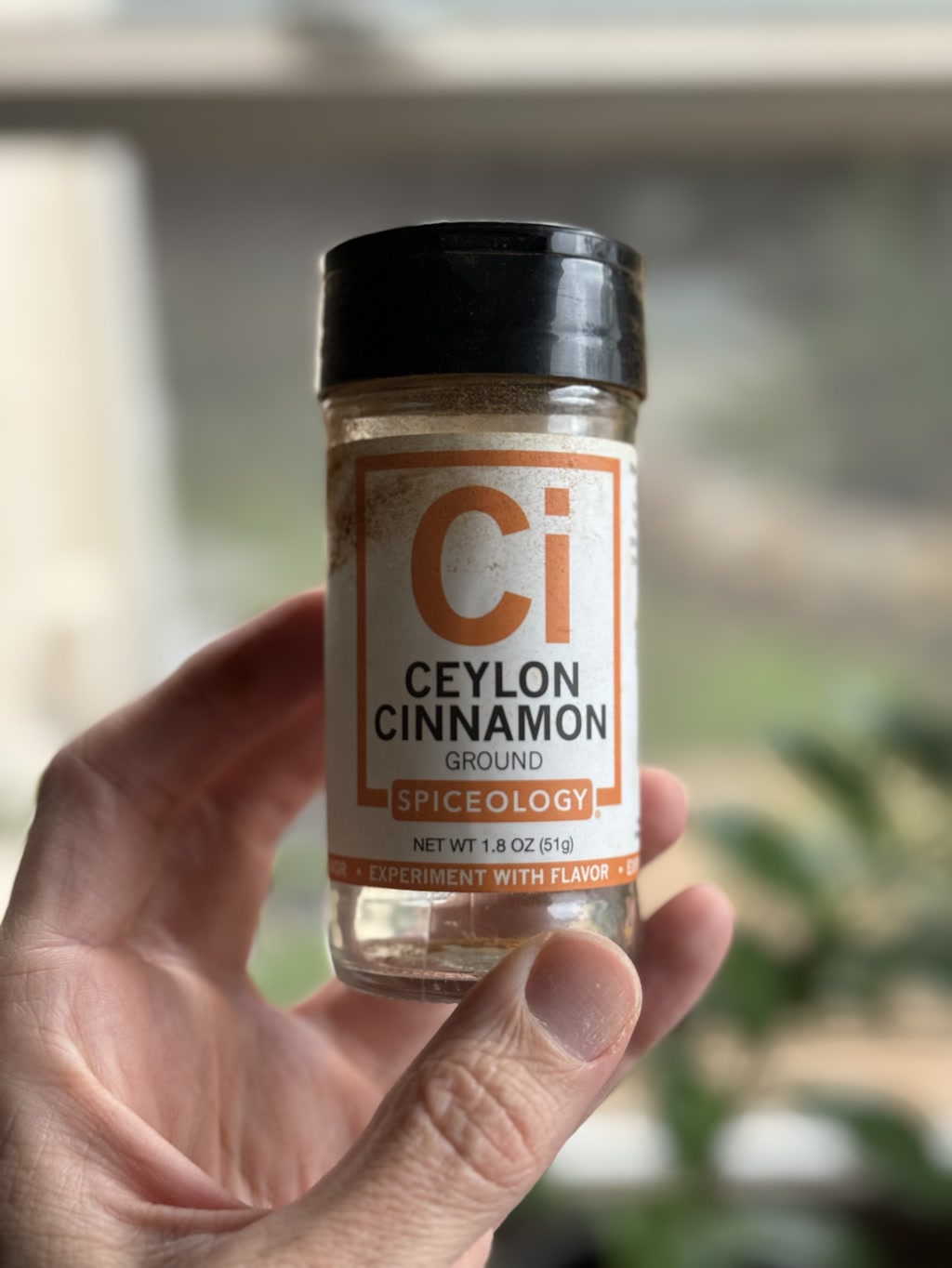 A hand is holding a jar of ground Ceylon Cinnamon by Spiceology. The label on the jar is predominantly white and orange with text providing product information. The jar has a black lid, and the contents are visible through the glass. In the background, there appears to be a blurred indoor environment with daylight coming through a window.